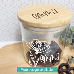 May include: A clear glass jar with a wooden lid, featuring the name "Emma" in a script font on both the lid and the jar. A white floral design is also on the jar. The jar contains dark-colored treats. The text "More designs available" is at the bottom.