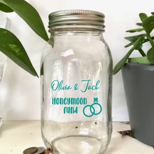 Pode incluir: Um frasco de vidro transparente com tampa prateada, etiquetado "Olivia & Jack Honeymoon Fund" com um design turquesa de dois anéis entrelaçados.
