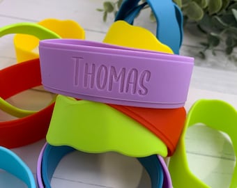Puede incluir: Un conjunto de pulseras de silicona de colores, algunas con un borde ondulado, con el nombre "THOMAS" grabado en una de las pulseras.