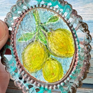 May include: An oval-shaped decorative piece featuring a hand-painted illustration of three yellow lemons on a textured blue background. The piece is encased in a scalloped, metallic frame with a weathered, oxidized teal and copper-toned finish.