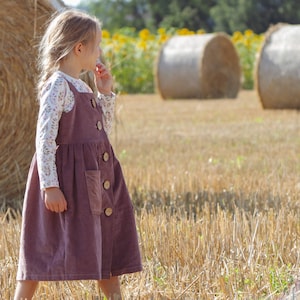 May include: A young girl wearing a brown corduroy dress with a floral print top and brown buttons. She is standing in a field of hay bales.