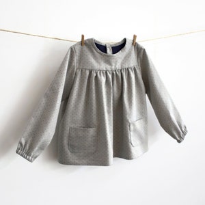 May include: A grey, long-sleeved blouse with a round neckline and a gathered bodice. The blouse has a polka dot pattern and two patch pockets.