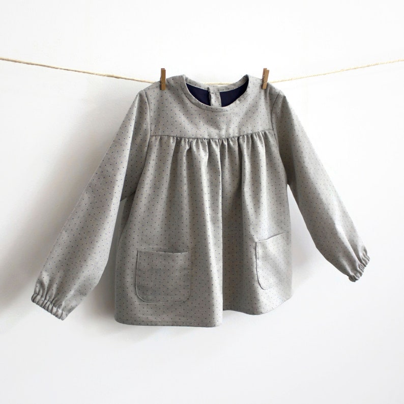 May include: A grey, long-sleeved blouse with a round neckline and a gathered bodice. The blouse has a polka dot pattern and two patch pockets.