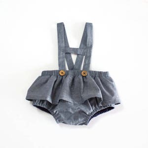 Peut inclure: Une barboteuse bébé en denim gris avec une jupe à volants et des bretelles. La barboteuse a deux boutons en bois sur le devant.