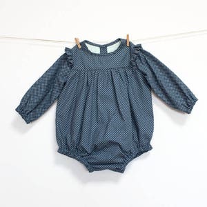 May include: A navy blue baby romper with white polka dots, long sleeves, and ruffled shoulders. The garment is hanging on a clothesline with wooden pegs. The romper has a round neckline and a gathered waist.