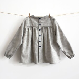 May include: A grey button-down shirt with a white polka dot pattern and long sleeves. The shirt has a gathered neckline and a button closure down the front.