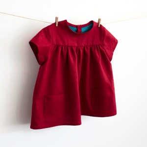 May include: A red short-sleeved dress with a gathered bodice and two pockets. The dress is hanging on a clothesline.