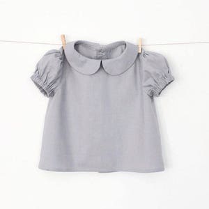 Puede incluir: Una blusa gris claro con cuello y mangas cortas abullonadas. La blusa tiene un cuello Peter Pan.