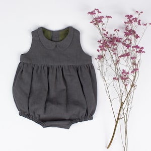 May include: A gray linen baby romper with a peter pan collar and a gathered skirt. The romper has a green lining around the collar.