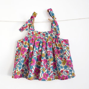 Pode incluir: Um vestido sem mangas de estampa floral branca com flores rosa, azuis e amarelas. O vestido tem duas alças que se amarram nos ombros.