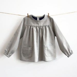 May include: A grey long-sleeved shirt with a white polka dot pattern. The shirt has a round neckline and two patch pockets on the front.