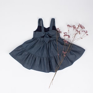 Puede incluir: Un vestido de mezclilla azul con un detalle de lazo en la espalda. El vestido tiene una falda de capas y es sin mangas.