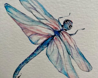 May include: Watercolor painting of a dragonfly with blue and pink wings. The dragonfly is in flight and its wings are spread out. The painting is signed "N.Robaina".