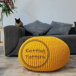 Op de afbeelding: Een ronde, gele gebreide poef met de tekst "Knitting Pattern" in een cirkel. De poef staat op een houten vloer voor een grijze bank met grijze kussens. Een kat zit op de bank.
