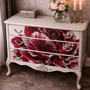 Könnte beinhalten: Weiß lackierte Kommode mit drei Schubladen, verziert mit einem großen rot-weißen Blumendesign. Die Kommode hat verzierte goldfarbene Griffe und dekorative Akzente. Eine Vase mit Blumen, Kerzen und eine dekorative Box stehen oben.
