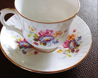 May include: A white teacup and saucer with a gold rim and floral pattern. The flowers are pink, yellow, blue, and green.