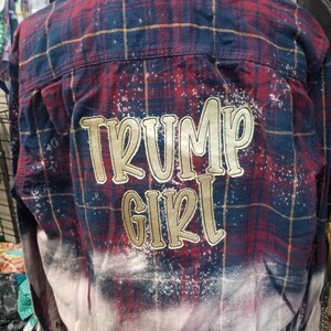 May include: A red, navy blue, and gold plaid shirt with the words "TRUMP GIRL" in gold lettering. The bottom of the shirt has a bleached, faded effect.