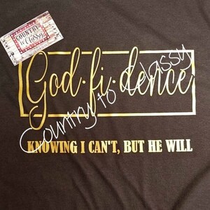 May include: Black t-shirt with gold script that reads "God-fidence" inside a gold rectangle. The phrase "Knowing I can't, but He will" is printed in gold below. A small tag with the text "Country to Classy" is in the upper left corner.