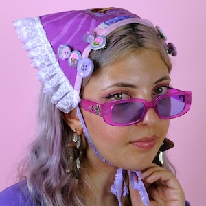 Puede incluir: Un pañuelo morado con borde de encaje blanco, decorado con varios botones morados y un pequeño parche rectangular. El pañuelo se ata bajo la barbilla con cintas de tela morada a juego. El accesorio se combina con unas gafas de sol de color rosa.