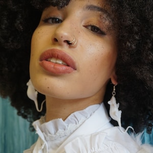 May include: A person with curly black hair wearing a white collared shirt with lace trim. They have a nose ring and earrings shaped like white mice.