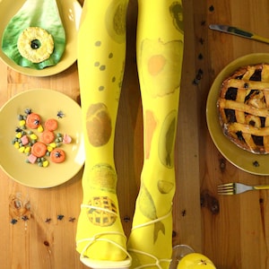 May include: Bright yellow patterned tights featuring fruit designs, worn with yellow sandals. The image includes plates with food items such as pie, pineapple, and other colourful treats, arranged on a wooden table.