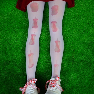 May include: White tights patterned with pink apple cores. The tights are worn with white trainers featuring red and white polka dot bows. The background is green.