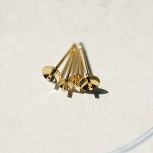 May include: Four small, gold-colored jewelry findings with a flat, circular top and a long, slender stem. The findings are arranged in a cluster, casting shadows on a light-colored surface. The overall aesthetic is minimalist and elegant.