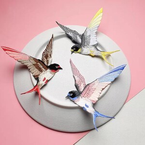 May include: Four embroidered swallows with outstretched wings are arranged on a white plate against a pink background. The birds feature detailed stitching in various colors, including red, yellow, blue, and brown, creating a vibrant and artistic display.