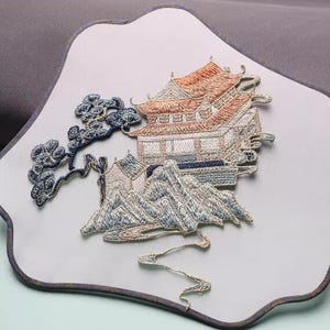 May include: Embroidered fan featuring a detailed landscape scene. The design includes a traditional building with an orange roof, mountains, and a tree branch, all rendered in shades of blue, white, and orange. The fan has a light gray background and a dark gray border.
