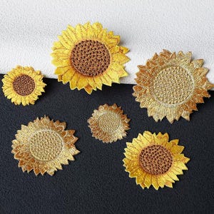 May include: A collection of six sunflower-shaped embroidered patches in varying sizes. The patches feature yellow petals and textured brown or gold centres, designed as decorative appliqués for fabric items with detailed stitching throughout.