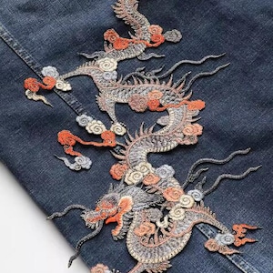 Puede incluir: Una chaqueta vaquera con dragones bordados en tonos grises, naranja y blanco. Los dragones están intrincadamente detallados y tienen un diseño chino tradicional.