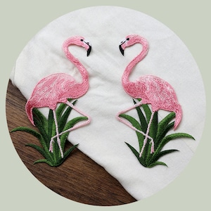 May include: Two pink flamingo embroidered patches with green leaves. The patches are on a white fabric background.