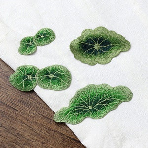 May include: Four green embroidered patches shaped like leaves. The patches are on a white fabric background.