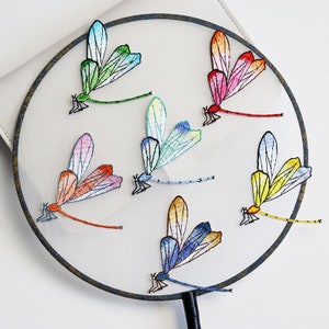 May include: A round, white hand fan with a dark brown border. The fan features embroidered dragonflies in various colors, including green, blue, yellow, orange, and pink. The dragonflies are arranged across the fan's surface.