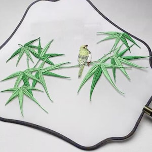May include: A white fabric fan with a black border and an embroidered design of a green bamboo branch with a small yellow bird perched on it.