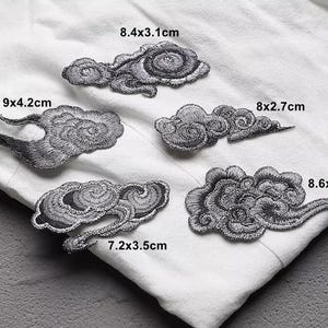 May include: Five embroidered cloud patches in various sizes, ranging from 7.2cm to 9cm in length, are displayed on a white fabric. The patches are grey with detailed stitching, showcasing a traditional design. The patches are ideal for sewing or crafting projects.