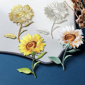 Peut inclure: Quatre écussons brodés en forme de tournesol avec des détails cousus. Un écusson est doré métallisé, un argenté métallisé, un jaune vif avec un cœur marron, et un blanc avec un cœur marron, tous dotés de tiges et de feuilles vertes.