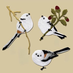Puede incluir: Parches bordados con aves blancas con marcas negras y naranjas. Dos aves se posan en ramas, mientras que una tercera descansa abajo. Los parches tienen costuras detalladas y están sobre un fondo neutro. Ideal para ropa o accesorios.