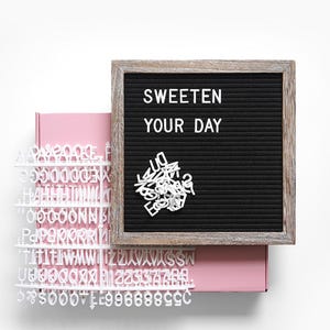May include: A black felt letter board with the words "SWEETEN YOUR DAY" in white letters. The board has a wooden frame and sits atop a pink box. Additional white letters and numbers are scattered around the board.