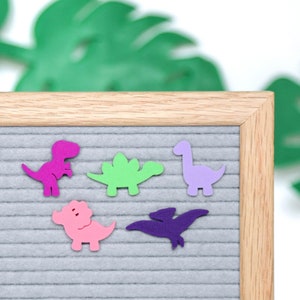 Puede incluir: Un tablero de letras de fieltro gris con un marco de madera, que presenta cinco formas de dinosaurios de fieltro: un estegosaurio rosa, un triceratops verde, un pterodáctilo morado, un tiranosaurio rex rosa y un braquiosaurio morado.