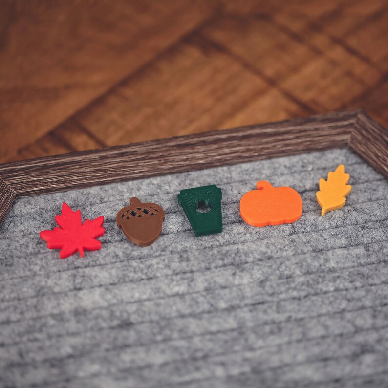 May include: Five felt fall-themed decorations on a gray background. The decorations are a red maple leaf, a brown acorn, a green coffee cup, an orange pumpkin, and a yellow leaf.