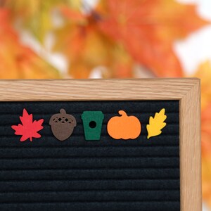 May include: A black felt letter board with a wooden frame. Five felt cutouts are attached to the board: a red maple leaf, a brown acorn, a green coffee cup, an orange pumpkin, and a yellow leaf.