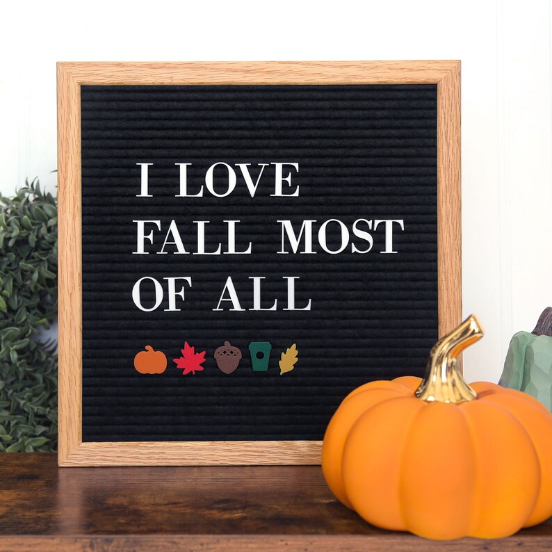 May include: A black letter board with the text "I love fall most of all" in white letters. There are small fall-themed decorations on the bottom row of the board, including a pumpkin, a leaf, an acorn, a coffee cup, and another leaf. A small orange pumpkin sits in front of the letter board.