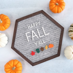 May include: A gray felt letter board with the text "Happy Fall Y'all" in white letters. The board is surrounded by small orange pumpkins and fall-themed decorations.