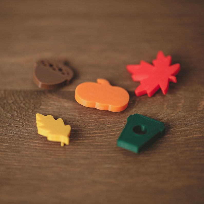 May include: Five small, colorful, 3D printed shapes on a wooden surface. The shapes include a brown acorn, an orange pumpkin, a yellow leaf, a red maple leaf, and a green coffee cup.