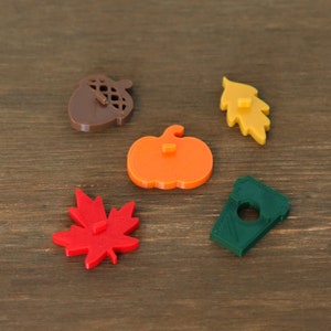 May include: Five colorful fall-themed shapes, including an acorn, a pumpkin, a maple leaf, an oak leaf, and a coffee cup, are arranged on a wooden surface.