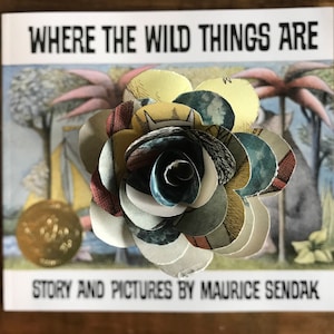 May include: A paper flower made from pages of the children's book "Where the Wild Things Are" by Maurice Sendak. The flower is multi-colored and has a blue center.