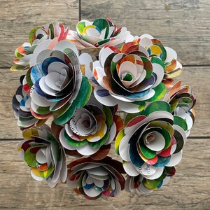 May include: A bouquet of paper flowers, each crafted from pages of a book. The flowers feature a layered design with visible text and vibrant colors including red, green, blue, and yellow. The arrangement is round and sits on a wooden surface.