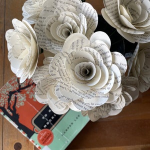 May include: A bouquet of white paper roses made from the pages of books. The roses are arranged in a vase and are sitting on a wooden table. A book titled "To Kill a Mockingbird" is visible in the foreground.