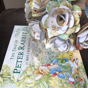 May include: A bouquet of paper flowers made from pages of the book "The Tale of Peter Rabbit" by Beatrix Potter. The flowers are white with green and brown accents. The book is visible in the background.